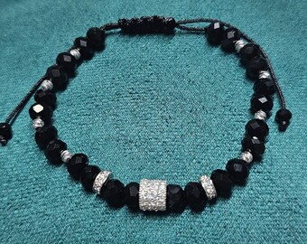 Peut inclure: Bracelet de perles noires avec des accents argentés et une perle rectangulaire centrale incrustée de petits cristaux étincelants. Le bracelet est réglable avec une fermeture à nœud coulissant, sur fond turquoise.