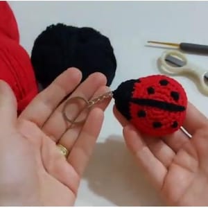 Op de afbeelding: Een handgemaakte gehaakte lieveheersbeestje sleutelhanger in rood en zwart, met zwarte stippen. De sleutelhanger wordt in de hand van een persoon gehouden, met rood en zwart garen en knutselgereedschap op de achtergrond.