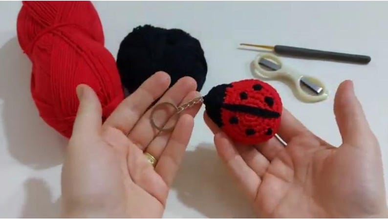 Op de afbeelding: Een handgemaakte gehaakte lieveheersbeestje sleutelhanger in rood en zwart, met zwarte stippen. De sleutelhanger wordt in de hand van een persoon gehouden, met rood en zwart garen en knutselgereedschap op de achtergrond.