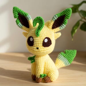 Può includere: Un peluche all'uncinetto fatto a mano a forma di personaggio dei cartoni animati. Il giocattolo è principalmente giallo con dettagli verdi e marroni. Ha grandi occhi rotondi, orecchie a punta e una coda a forma di foglia. Il giocattolo è su una superficie di legno.