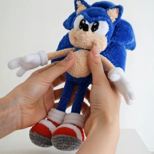 Può includere: Bambola di Sonic the Hedgehog all'uncinetto fatta a mano, blu, bianca, rossa e grigia. La bambola ha guanti bianchi, scarpe rosse e bianche e una pancia color beige. La bambola è tenuta da due mani.