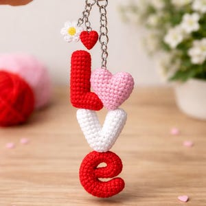 May include: A handmade crochet keychain spelling "LOVE" in red and white yarn. The "O" is red, the "V" is white, and the "L" and the heart-shaped "E" are red. A small daisy and a red heart charm dangle from the keychain.