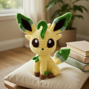 May include: A handmade crochet plush toy in the shape of a cartoon animal. The toy is primarily yellow with green accents on the ears, tail, and small leaves. It has brown eyes and is sitting on a cream-colored pillow.