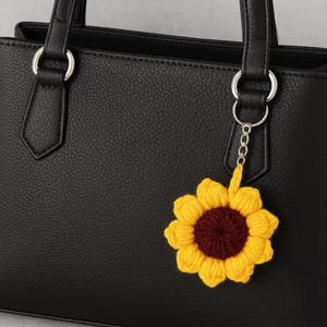Puede incluir: Bolso de mano de cuero negro con aros plateados y un llavero de girasol tejido a ganchillo. El girasol tiene pétalos amarillos y un centro granate, añadiendo un toque de color.