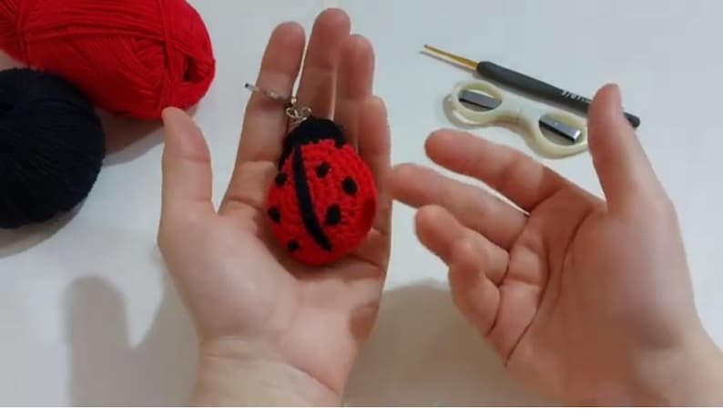 Op de afbeelding: Een handgemaakte rode en zwarte lieveheersbeestje sleutelhanger. Het lieveheersbeestje is gehaakt met zwarte stippen en een zwarte kop. Rood en zwart garen zijn zichtbaar op de achtergrond, samen met een haaknaald en een schaar.