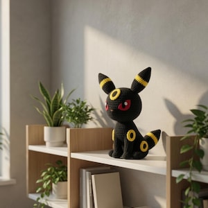 Puede incluir: Una figura de Umbreon negra de ganchillo con detalles amarillos y ojos rojos está colocada en una estantería blanca. La figura tiene orejas grandes y una cola con anillos amarillos. La estantería forma parte de una estantería de madera con plantas y libros.