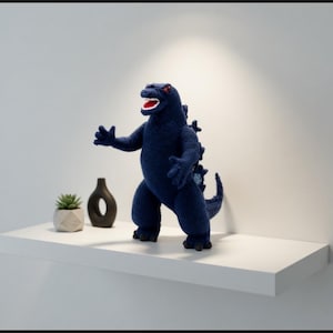 Könnte beinhalten: Eine marineblaue Godzilla-Figur aus Häkelarbeit mit roten Augen und einem weißen Mund steht auf einem weißen Regal. Eine kleine Sukkulente in einem weißen Topf und eine schwarze Vase befinden sich ebenfalls auf dem Regal. Der Hintergrund ist eine schlichte weiße Wand.