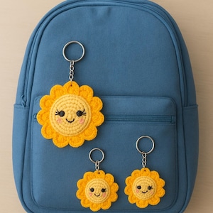 Puede incluir: Una mochila azul con tres llaveros de sol de ganchillo amarillos. Los soles tienen caras sonrientes y están unidos a llaveros plateados. Los llaveros son de diferentes tamaños. La mochila tiene un bolsillo frontal.