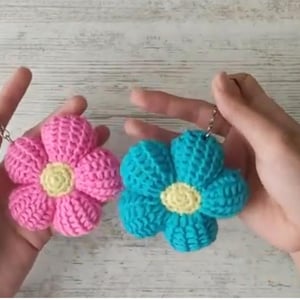 Könnte beinhalten: Zwei handgefertigte Schlüsselanhänger in Form von gehäkelten Blumen. Einer ist rosa mit gelber Mitte, der andere türkis mit gelber Mitte. Beide haben silberfarbene Schlüsselringe. Die Blumen werden vor einem hellen Holzhintergrund gehalten.