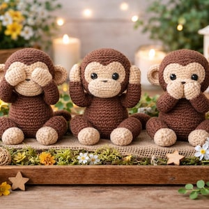 Puede incluir: Tres monos de ganchillo hechos a mano en la pose 'No ver el mal, no oír el mal, no hablar el mal'. Los monos marrones tienen caras y pies beige, sentados en una bandeja de madera con flores y vegetación. Un artículo decorativo.