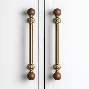 May include: Two cabinet pulls with a brass finish and wooden ball finials. The pulls are mounted on a white cabinet door. The design features a vertical bar with decorative elements.