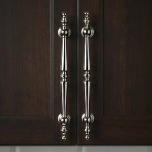 May include: Two polished nickel cabinet pulls with a decorative design. The pulls are vertically mounted on a dark brown cabinet door. The cabinet door has a smooth, wood grain finish. The pulls are symmetrical and have a classic, elegant style.