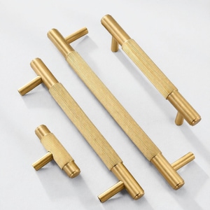 May include: Four gold cabinet pulls with a ribbed texture and T-shaped handles. The pulls vary in length, with the longest measuring approximately 30 cm and the shortest about 10 cm. They are arranged on a white surface.