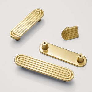 May include: A selection of gold-toned cabinet handles and a knob. The handles have a grooved design. The hardware is displayed on a white background. The longest handle is approximately 15 cm long.