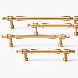 May include: Five gold-coloured cabinet pulls are displayed. Each pull has a central, gently curved bar with decorative, rounded ends and vertical posts for mounting. The pulls are arranged against a white background.