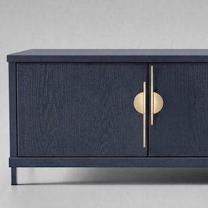 May include: A navy blue cabinet with two doors and gold-coloured hardware. The cabinet has a rectangular shape and is supported by thin, dark legs. The doors feature a wood grain texture.
