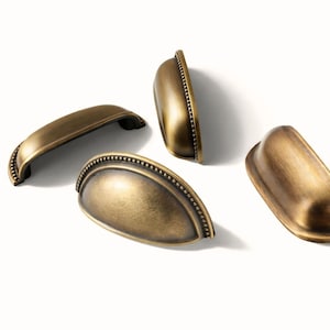 Puede incluir: Cuatro tiradores de armario de latón antiguo en varias orientaciones. Los tiradores tienen un diseño curvo con un detalle de borde con cuentas. El acabado es un color latón cálido y envejecido, lo que sugiere una estética vintage o rústica.