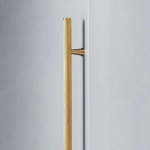 Puede incluir: Un tirador de puerta dorado y texturizado. El tirador es cilíndrico con un patrón de puntos y está montado sobre una superficie blanca. El tirador es una barra vertical con dos puntos de montaje.