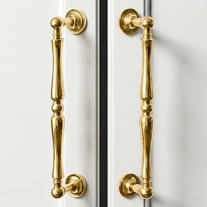 May include: Two gold-colored cabinet pulls with a classic design are mounted on a white cabinet door. The pulls have a vertical, elongated shape with decorative detailing and rounded ends. The hardware adds a touch of elegance.