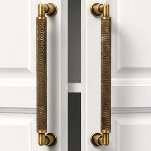 May include: Two antique brass cabinet pulls with a textured, cylindrical grip. The pulls are mounted on a white cabinet door, showcasing a classic design. The hardware adds a touch of vintage charm to any furniture piece.
