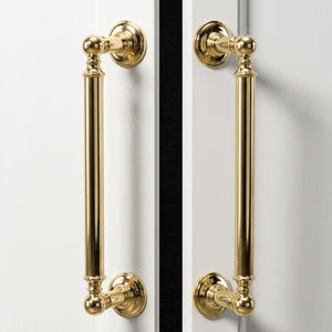 May include: Two polished gold door handles. Each handle features a long, cylindrical grip with decorative ball-shaped accents at the top and bottom, and a circular base. The handles are mounted on a white door.