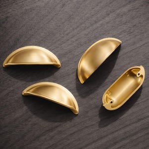 Può includere: Quattro maniglie dorate per mobili a forma di mezza luna sono disposte su una superficie in legno scuro. Tre sono curve, mentre una ha la forma di una tazza rettangolare con fori per le viti.