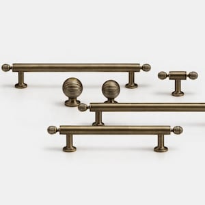 May include: Collection of antique brass cabinet hardware, including various sizes of pulls and knobs. The pulls have a bar design with decorative ball ends. The knobs have a ribbed, spherical design. The hardware is displayed against a white background.