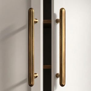 May include: Close-up of white cabinet doors with long, gold-colored handles. The handles have a textured, dotted pattern and are mounted vertically. The cabinet doors are slightly ajar, revealing a glimpse of the interior.