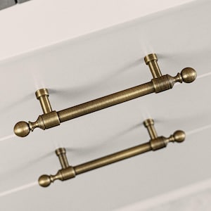 May include: Two antique brass cabinet pulls. Each pull has a cylindrical bar with decorative ball finials. The pulls are mounted on a light grey surface, possibly a drawer or cabinet door. The pulls are made of brass.