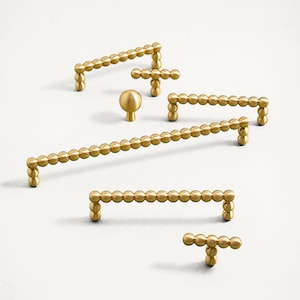 May include: A selection of gold-coloured cabinet hardware, including knobs and pulls. The hardware features a beaded design, with small, round beads forming the handles and knobs. The pieces are arranged on a white background, showcasing their design.