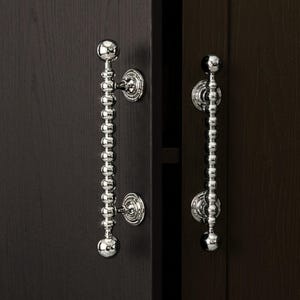 Puede incluir: Primer plano de dos puertas de armario con tiradores plateados pulidos. Cada tirador presenta una serie de pequeñas cuentas esféricas, con una esfera más grande en cada extremo. Montados en un armario de madera marrón oscuro.