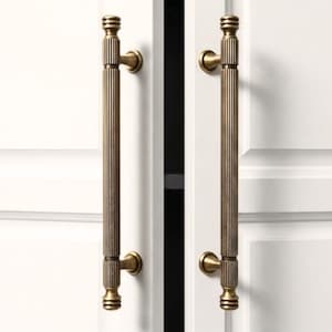 Peut inclure: Deux poignées de placard en laiton antique avec un design côtelé vertical. Les poignées sont montées sur une porte d'armoire blanche. Les poignées ont une forme cylindrique avec des détails décoratifs en haut et en bas.