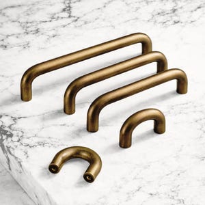 May include: A selection of bronze-toned cabinet handles in different sizes. The curved, tubular handles are displayed on a white marble surface. These handles are designed for use on drawers and cabinets, and are measured in centimetres.