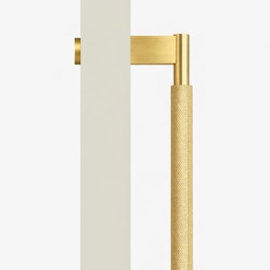 May include: A brass-coloured door pull handle with a textured, cylindrical grip. The handle is attached to a cream-coloured door panel. The design features a rectangular base and a vertical bar with a dotted pattern for grip.