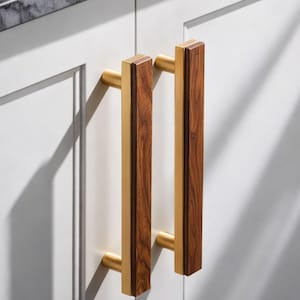 May include: Two cabinet pulls with a rectangular design. The pulls feature a combination of wood and gold-toned metal. The wood is a medium brown color, and the metal has a shiny finish. The pulls are mounted on a white cabinet door.