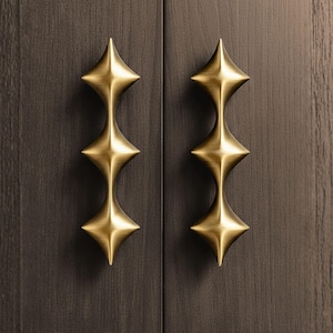 May include: Two brass cabinet pulls with a starburst design are mounted on a dark wood cabinet door. The pulls have a three-tiered, diamond-shaped design, adding a touch of elegance to the furniture.