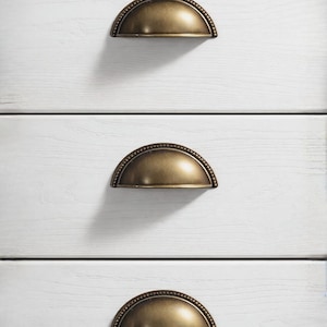 Puede incluir: Primer plano de una cómoda blanca con tres cajones. Cada cajón tiene un tirador en forma de media luna de color bronce con un borde con cuentas. La veta de la madera es visible en la superficie pintada de blanco.