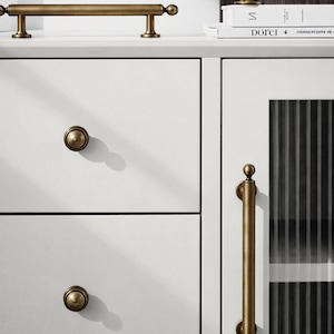 May include: Close-up of white cabinetry with brass hardware. The image features a long bar handle, round knobs, and a vertical pull handle. A decorative brass sphere sits atop a stack of books.