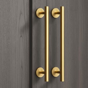 Peut inclure: Deux poignées de placard dorées avec une barre cylindrique. Chaque poignée a une base circulaire avec des rainures concentriques. Les poignées sont montées sur une porte de placard en bois gris foncé.