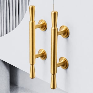 May include: Two gold-colored cabinet pulls are mounted on a white surface. Each pull has a cylindrical design with a flared base and top, and is attached to the surface with a circular base. The pulls are vertically oriented.