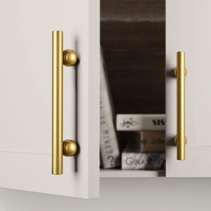 May include: Two white cabinet doors with gold-coloured bar handles. The handles are cylindrical with a spherical accent at each end. One door is slightly ajar, revealing a glimpse of books inside the cabinet.