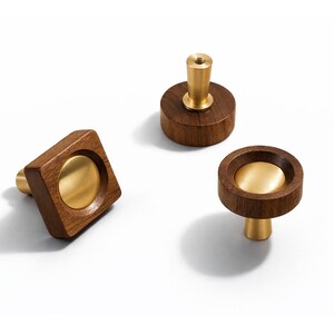 May include: Three wooden cabinet knobs with brass accents. One is square, one is round, and one is cylindrical. The knobs are made of dark wood and have a modern design. The brass hardware adds a touch of elegance.