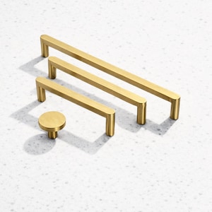 May include: A collection of gold-coloured cabinet hardware, featuring two rectangular bar pulls and a round knob. The hardware is displayed on a white speckled surface, with shadows. The pulls are of different lengths, suitable for various cabinet sizes.