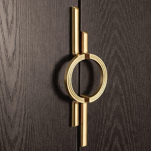 May include: A gold-colored cabinet pull with a circular design and vertical bars. The pull is mounted on a dark brown wooden cabinet door, showcasing a modern and elegant aesthetic. The hardware is made of metal.