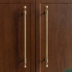 May include: Close-up of two wooden cabinet doors with long, brass-colored handles. The handles have a decorative, vertical design with a ball at the top and bottom. The wood grain is visible in the dark brown doors.
