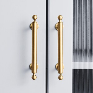 May include: Two gold-colored cabinet pulls with a cylindrical design and rounded ends. The pulls are mounted on a light gray cabinet door. The background includes a section of a ribbed glass panel.