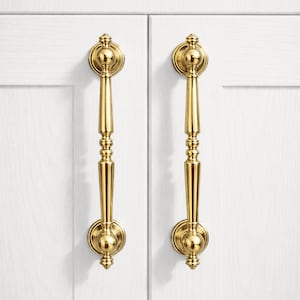 May include: Two polished brass cabinet pulls with a classic design. Each pull features a round backplate, a decorative center, and a long, elegant handle. The pulls are mounted on a white cabinet door.
