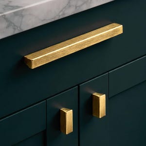 May include: Close-up of a dark teal cabinet with gold-coloured hardware. The cabinet has a long rectangular handle and two smaller square knobs. A white and grey marble countertop is visible in the background.
