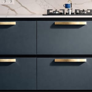 May include: A modern kitchen cabinet design featuring navy blue cabinet doors with gold-toned rectangular handles. The countertop is white with a marble pattern. A stovetop and a blue coffee pot are visible.
