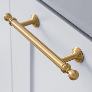 May include: A gold-coloured cabinet pull with a cylindrical bar and decorative ball finials. The hardware is mounted on a white cabinet door, offering a classic and elegant design for kitchen or bathroom cabinetry.